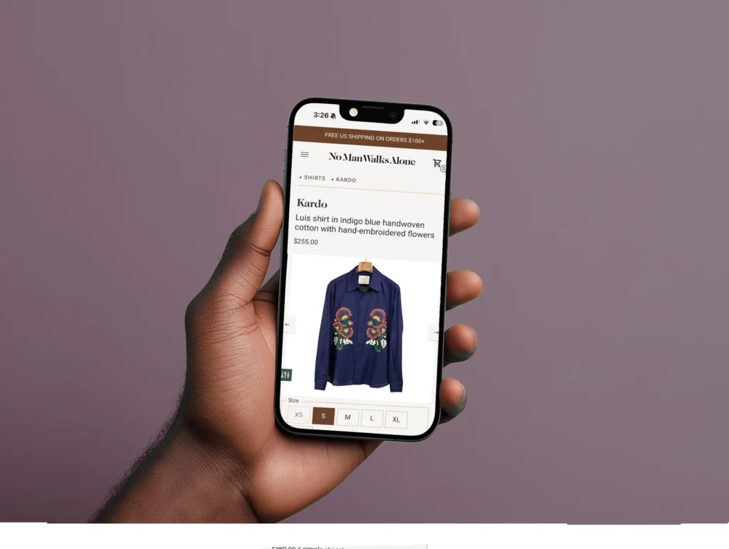 Hand holding a smartphone displaying a product page for a 'Luis shirt in indigo blue handwoven cotton with hand-embroidered flowers' on the No Man Walks Alone website, priced at $255. The page includes product details such as size options (XS, S, M, L, XL).