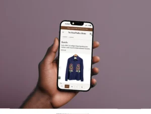 Hand holding a smartphone displaying a product page for a 'Luis shirt in indigo blue handwoven cotton with hand-embroidered flowers' on the No Man Walks Alone website, priced at $255. The page includes product details such as size options (XS, S, M, L, XL).