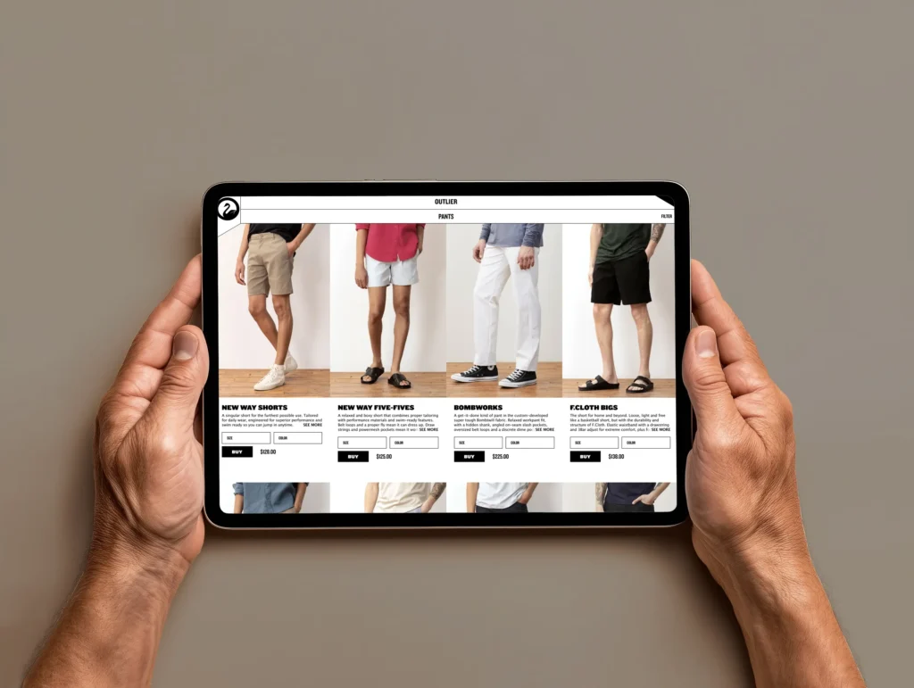 Hands holding a tablet displaying the Outlier product page for shorts, showcasing different styles including New Way Shorts, New Way Five-Fives, Bombworks, and Fcloth Bigs.
