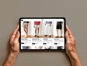 Hands holding a tablet displaying the Outlier product page for shorts, showcasing different styles including New Way Shorts, New Way Five-Fives, Bombworks, and Fcloth Bigs.