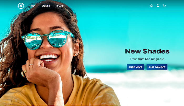 Woman sitting on the beach and smiling. She wears sunglasses the color of tropical blue sky