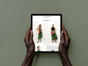 Hands holding a tablet displaying the ‘New Arrivals’ section of a fashion website, featuring a model wearing a vibrant green and red floral dress, set against a clean white background on the product page.