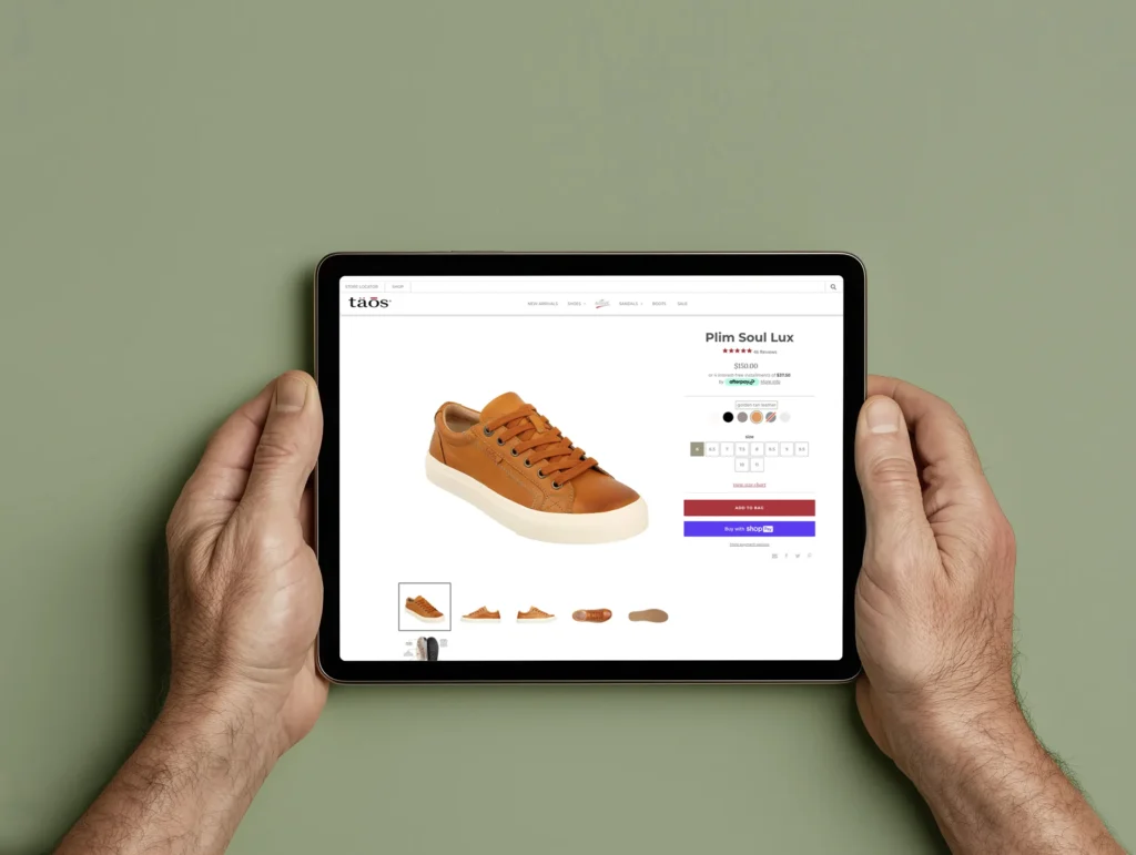 Person holding a tablet displaying the product page for Taos Footwear's Plim Soul Lux sneaker in golden tan leather, showing product details, color options, size selection, and the 'Add to Bag' button on the ecommerce website.
