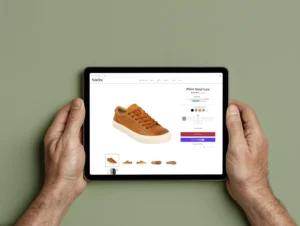 Person holding a tablet displaying the product page for Taos Footwear's Plim Soul Lux sneaker in golden tan leather, showing product details, color options, size selection, and the 'Add to Bag' button on the ecommerce website.