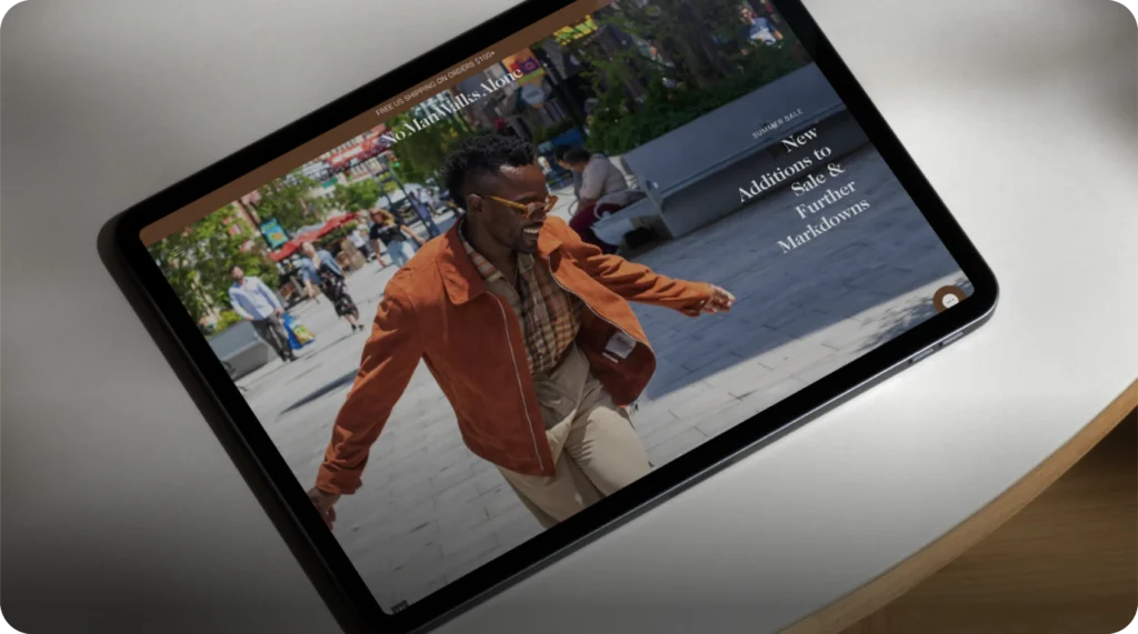 Tablet displaying a fashion website with a man in a tan suede jacket and beige pants walking joyfully outdoors. The screen highlights a summer sale promotion with the text 'New Additions to Sale & Further Markdowns.